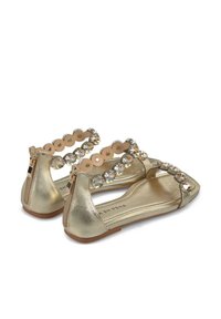 Gouden flats met een glanzende metallic afwerking, voorzien van een decoratieve band met ronde en edelsteenaccenten, en een ritssluiting aan de achterkant.