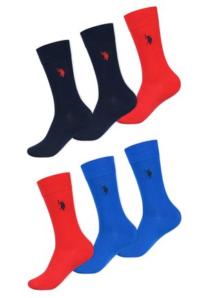 Mann trägt schwarze Shorts, marineblaue knielange Socken und ein weißes Hemd und geht vor einem einfarbigen hellen Hintergrund.