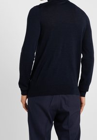 Námorně modrý vlněný svetr s dlouhými rukávy, kulatým výstřihem a žebrovaným lemem. Hladká textura s minimálním zdobením, nošený přes tmavé kalhoty.