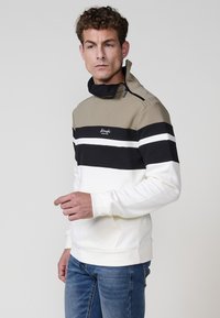 Polovica zips pulóver v béžovej, čiernej a bielej prúžkovej kombinácii. Vyrobený z mäkkej tkaniny, má vysoký golier, logo značky na hrudi a prednú kapsu.