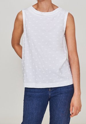 Kvinde iført hvid tekstureret ærmeløs top med diskret polkaprikket mønster og blå denimjeans, stående med den ene hånd bag ryggen.