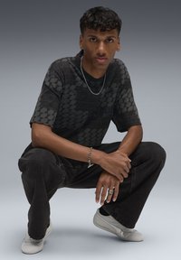 Czarny graficzny t-shirt z sześciokątnym wzorem, czarne dżinsy i jasnoszare trampki. Akcesoria obejmują srebrny naszyjnik z łańcuszka oraz pierścionki.