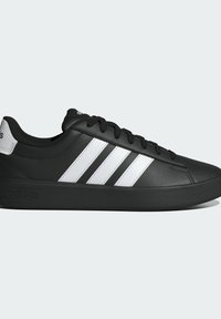 Baskets Adidas basses noires avec trois bandes blanches et lacets, logo en relief sur la semelle et languette blanche au talon sur fond neutre.