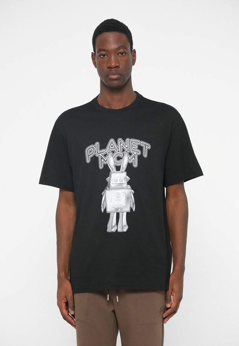 Muškarac u crnoj majici s natpisom "PLANET MCM" i grafikom srebrnog robota zeca, u kombinaciji s smešnim hlačama, stoji naslonjen na bijelu pozadinu.