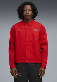 Jachetă roșie cu închidere frontală cu nasturi, având guler și logo-ul brodat "Scuderia Ferrari" în gri. Textură de țesătură netedă.