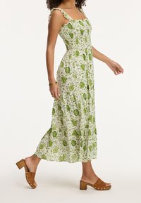 Grünes Maxikleid mit floralem Muster, Rüsche-Trägern, gerafftem Oberteil und einem fließenden Rock. Kombiniert mit braunen Clogs mit Blockabsatz.