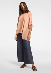 Hedvábný top v broskvové barvě se splývavými rukávy, vrstveným předním dílem, kombinovaný s kalhotami z tmavého denimu s širokými nohavicemi a černými plochými sandály.