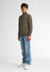 Maglione a collo alto verde oliva, lavorato a maglia media, abbinato a jeans azzurri chiari e sneaker bianche. Design semplice, silhouette aderente, senza motivi.