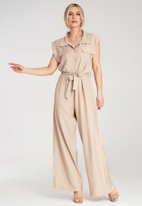 Beżowy kombinezon z szerokimi nogawkami, krótkimi rękawami, zapięciem na guziki z przodu i przewiązanym w pasie. Gładki materiał o luźnym fasonie i dwiema przednimi kieszeniami.