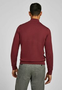 Mann in einem figurbetonten kastanienbraunen Pullover und grauen strukturierten Stoffhosen, von hinten vor einem schlichten hellen Hintergrund gezeigt.