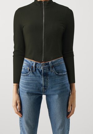 Personne portant un haut ajusté, côtelé, vert foncé avec fermeture éclair et un jean bleu taille haute, debout devant un fond uni.