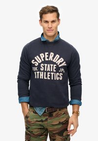 Marineblauwe sweatshirt met witte tekst "SUPERDRY TOK. STATE JPN ATHLETICS," gedragen over een lichtblauw denim shirt en camouflagebroek.