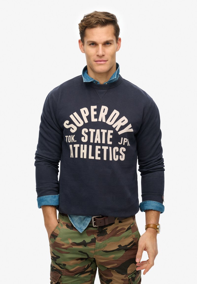 Marineblauwe sweatshirt met witte tekst "SUPERDRY TOK. STATE JPN ATHLETICS," gedragen over een lichtblauw denim shirt en camouflagebroek.
