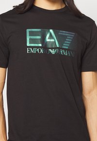 Svart bomull T-shirt med en glänsande turkos EMPORIO ARMANI-logo med stiliserade bokstäver och horisontella linjer över designen.