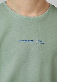 Lichtgroen shirt met blauwe tekst die zegt "ENKEL EINDLOZE MOMENTEN VERZAMELEN ESCAPE NAAR HET PARADIJS Sunset STATE OF LIME" dicht bij de kraag.
