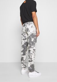 Ein Mann trägt ein schwarzes T-Shirt und eine weiß-graue Batik-Cargohose, steht seitlich mit weißen Turnschuhen vor einem schlichten Hintergrund.