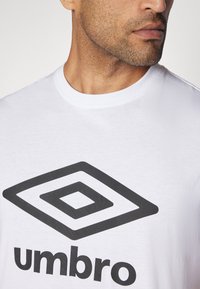 Hvid bomulds t-shirt med et stort sort diamantlogo og ordet "umbro" i fed skrift under grafikken. Glat tekstur, rund halsudskæring.