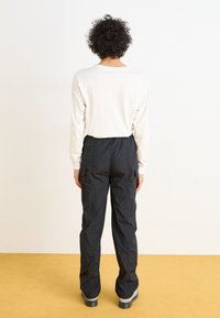 Pantalons cargo noirs avec taille élastique et poches, associés à une chemise blanche à manches longues, vus de dos. Chaussettes et chaussures visibles.