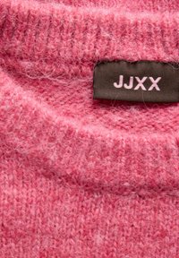 Gros plan d'un tissu tricoté rose doux et duveteux avec une étiquette noire brodée des lettres JJXX en fil rose clair.
