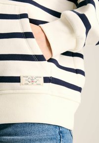 Biely a námornícky pruhovaný svetrov s prednou vreckom a rebrovanými manžetami. Obsahuje značkovú nálepku na dolnej časti predného dielu. Mäkká textúra látky.