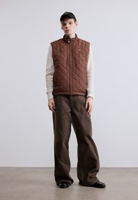 Καφέ καπιτονέ γιλέκο με ψηλό γιακά, φορεμένο πάνω από ένα ανοιχτόχρωμο, υφασμάτινο πουλόβερ. Συνδυασμένο με άνετο καφέ παντελόνι και μαύρα παπούτσια. Μαύρη καπέλο στο κεφάλι.