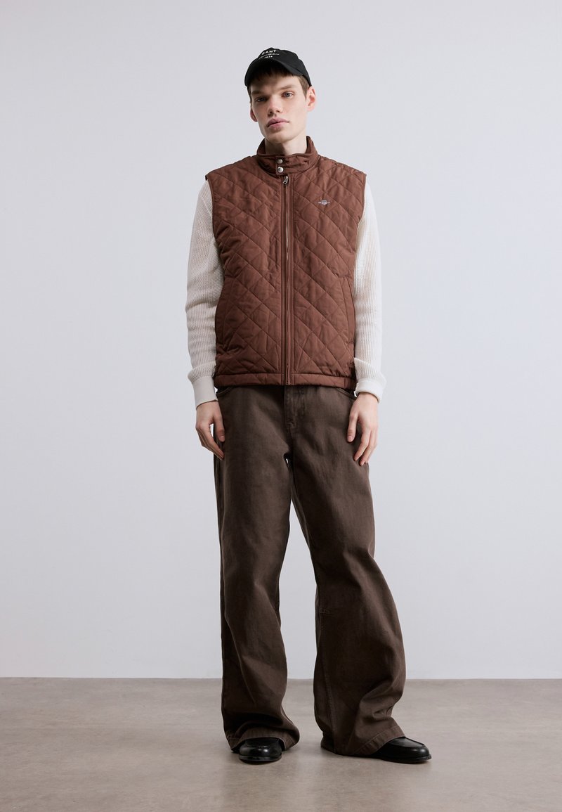 Brown quilted vest with a high collar, worn over a light, textured sweater. Paired with loose brown trousers and black shoes. Black cap on head.