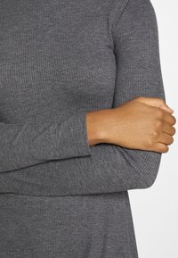 Haut à manches longues côtelé gris avec une texture douce, présentant une coupe ajustée et un col rond. Les extrémités des manches dépassent légèrement le poignet.