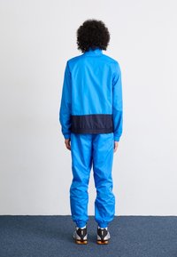 Survêtement bleu avec une bande marine à la taille. Le matériau est lisse et brillant, avec des poignets élastiques et des chevilles élastiques. Baskets visibles.