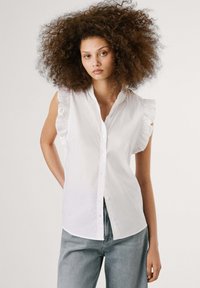 White sleeveless blouse with ruffled shoulder accents, button front, and a slightly curved hem. Made of smooth, lightweight fabric.