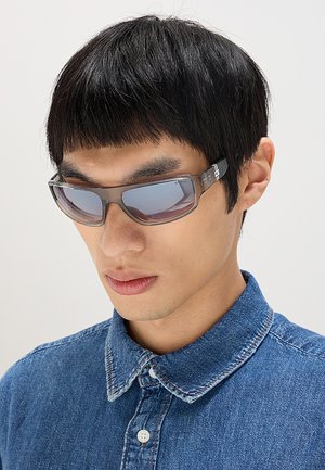 Giovane uomo con capelli neri e lisci, che indossa occhiali da sole rettangolari con lenti scure e una camicia di jeans blu con bottoni, guarda verso il basso.