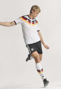 Jeune joueur de football masculin en tenue de l'équipe d'Allemagne, en train de frapper le ballon, portant un équipement blanc, noir, rouge et jaune avec des crampons et des chaussettes hautes.