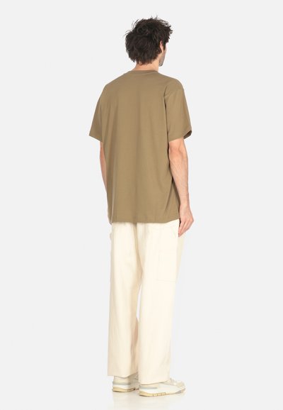 Uomo in piedi di spalle, vestito con una maglietta marrone oversize, pantaloni beige larghi con tasche laterali e sneakers bianche su uno sfondo bianco semplice.