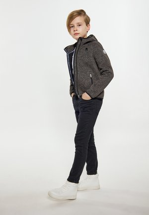Grijze zip-up jas met een capuchon, voorzien van twee voorzakken, gedragen over een zwart shirt en zwarte skinny jeans. Afgerond met witte sneakers.