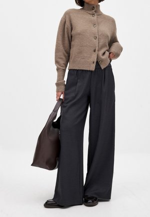 Femme portant un cardigan taupe boutonné, un pantalon large gris foncé, des chaussures noires, et tenant un grand sac cabas marron foncé.