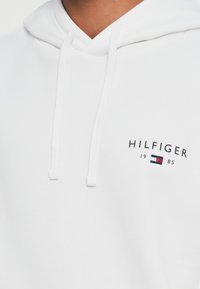 Sweatshirt branco em algodão, com capuz ajustável e um logótipo no peito com "HILFIGER" e "1985" em preto, realçado por um pequeno emblema em vermelho e azul.