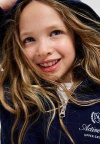 Sweat à capuche zippé marine avec une fermeture éclair argentée, arborant un texte blanc et un logo sur la poitrine. Tissu doux et manches longues, encadré par des cheveux blonds lâches.