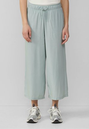 Pantalon court ample bleu clair avec taille à cordon, porté avec des baskets argentées et blanches par une personne debout devant un fond uni.