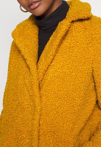 Cappotto giallo, con una texture soffice e pelosa, dotato di colletto a tacca, che presenta una superficie morbida al tatto, indossato sopra un indumento a collo alto nero.