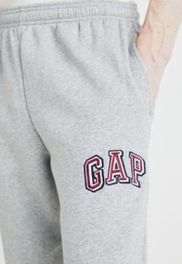 Sivé tepláky s elastickým pásom, bočnou vreckom a vyšitým logom "GAP" v červenej a modrej farbe na stehne. Mäkká, textúrovaná látka.