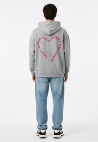 Felpa grigia con cappuccio e grande testo rosso a forma di cuore sulla schiena. Abbinata a jeans azzurri chiari e sneakers nere e bianche.