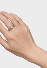 Anillo de malla plateada con un acento de trébol dorado. El anillo tiene un diseño flexible con una apariencia minimalista y una textura suave.