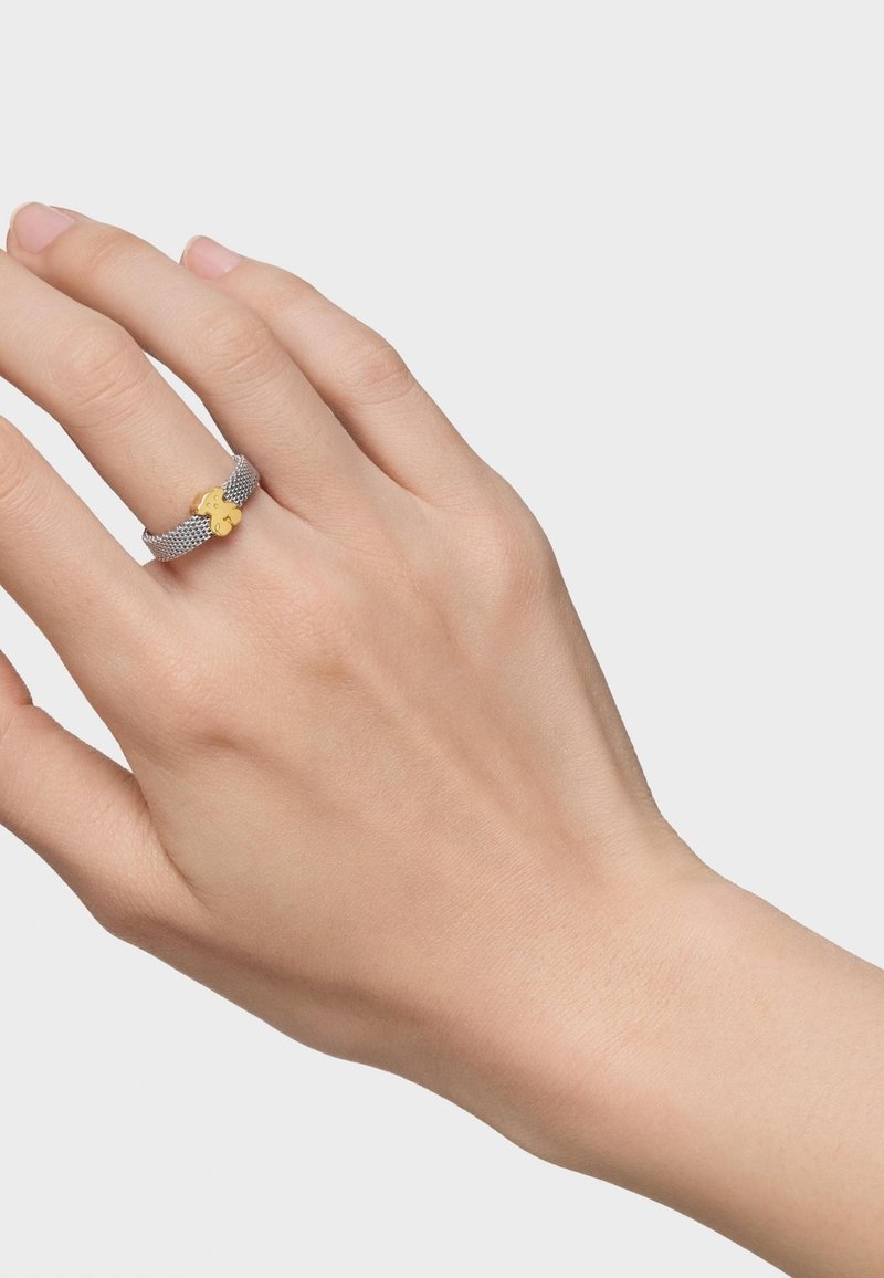 Anillo de malla plateada con un acento de trébol dorado. El anillo tiene un diseño flexible con una apariencia minimalista y una textura suave.