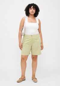 Pantalones cortos verde claro hechos de tela texturada, con un cierre de botón único y un dobladillo enrollado. Combinados con una blusa blanca de escote cuadrado y zapatos beige.