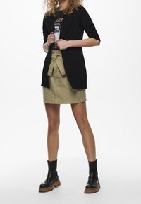 Cardigan texturé noir porté sur un t-shirt graphique noir, associé à une jupe kaki avec une taille nouée. Bottines noires avec semelles marron.