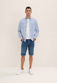 Vaaleansininen pitkähihaisen napillinen paita valkoisen T-paidan päällä, yhdistettynä sinisiin farkkushortseihin ja valkoisiin lenkkareihin. Kaksi rintataskua näkyvissä.