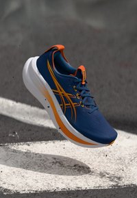 Blauer Sportschuh mit orangefarbenen Akzenten, strukturiertem Obermaterial, Schnürsenkeln und einer dicken weißen Sohle mit einem Verlaufdesign.