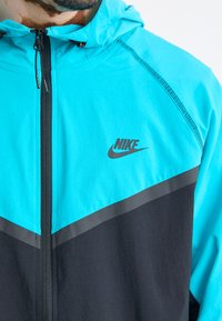Nike-jakke med en lys turkise toppseksjon, svart bunnseksjon, glidelås foran og logoakkent på brystet. Holdbart stoff med hette.