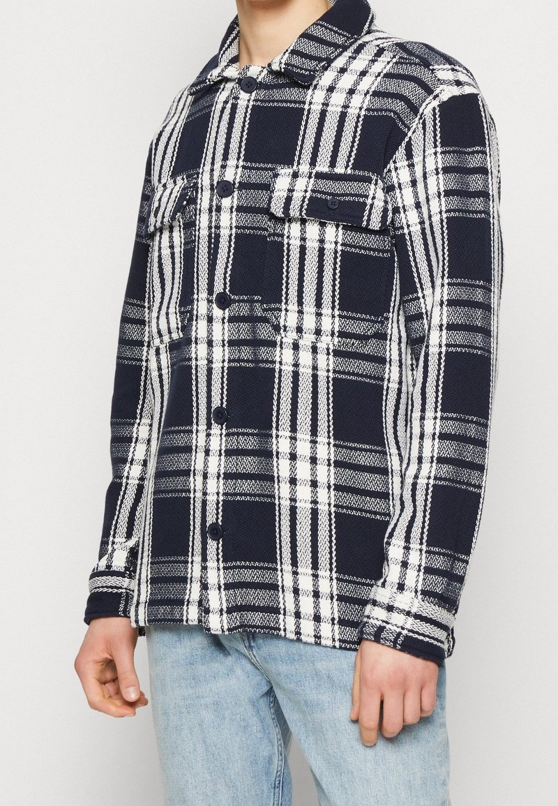 Mann trägt ein marineblau-weiß kariertes Hemd mit Knöpfen, aufgesetzten Brusttaschen mit Klappen und hellblaue Jeans vor einfachem Hintergrund.