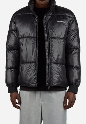 Mężczyzna ubrany w błyszczącą czarną pikowaną kurtkę z logo "PEGADOR", czarną koszulę pod spodem oraz jasnoszare spodnie z haftowanymi detalami.