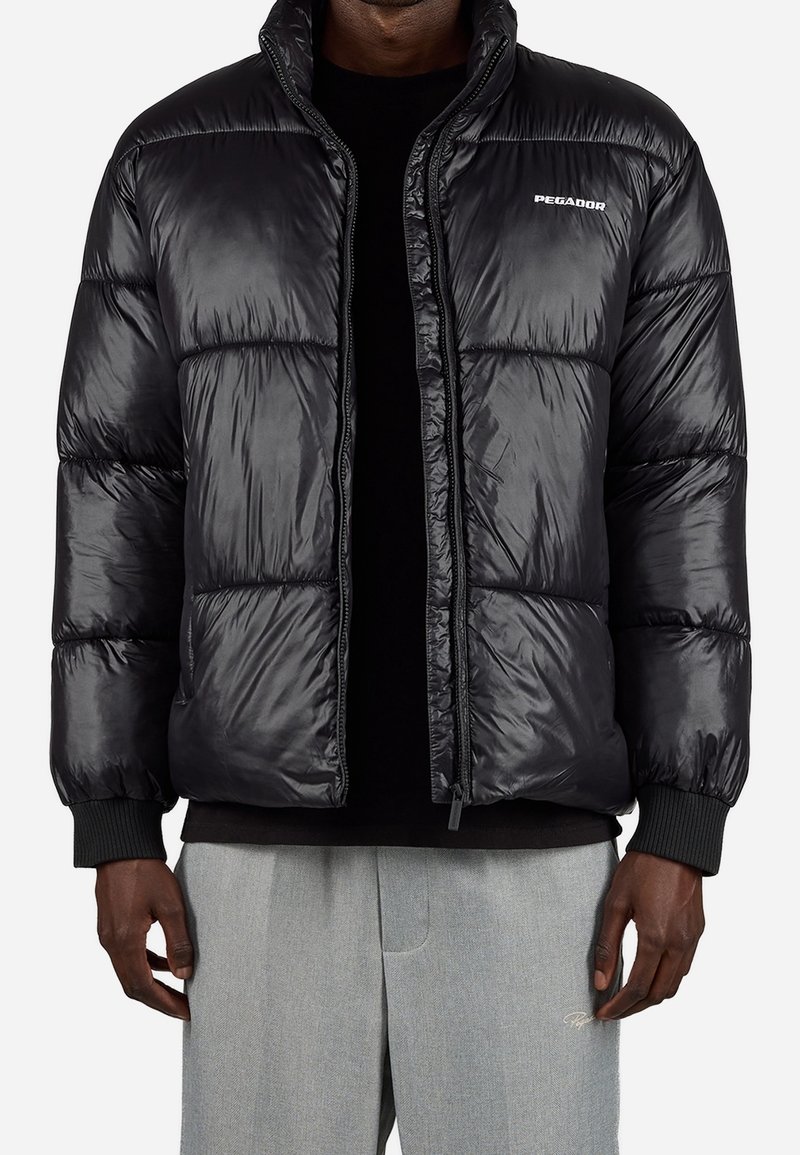 Homme portant une doudoune noire brillante avec le logo "PEGADOR", une chemise noire en dessous et un pantalon gris clair avec un détail brodé.
